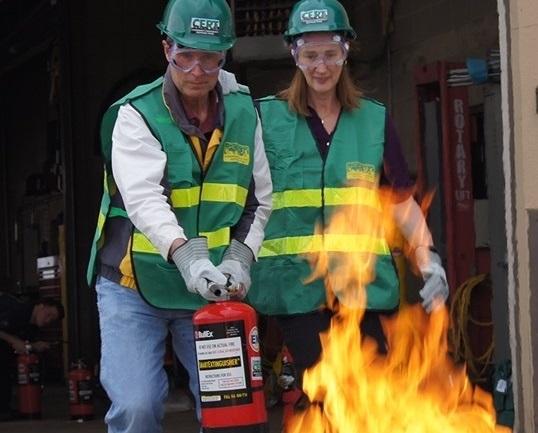 A man and woman wearing CERT green hard hats a green vests with two reflective safety stripes practice using a fire extinguisher on a small fire.