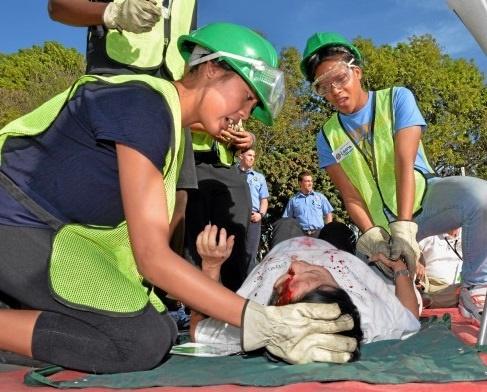Several CERT trainees wearing safety gear give a mock emergency first aid demonstration with a male volunteer "victim," who is lying face up on the ground. One rescuer is talking to the victim while a second is holding his hand. He has fake blood on his face.