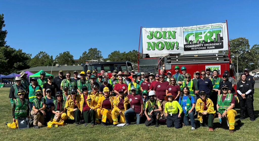 Over 50 CERT volunteers pose for a group photo in front of the CERT firetruck. Some wear green reflective vests or yellow reflective pants and jackets. Over and attached to the firetruck is a wide banner stretched between two poles. The banner has the words, "Join now!" on one side and the Northern Sonoma County CERT logo on the other.